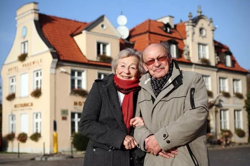 Starszy mężczyzna i kobieta stoją ubrani ciepło i przytuleni do siebie, w tle panorama Gdańska 