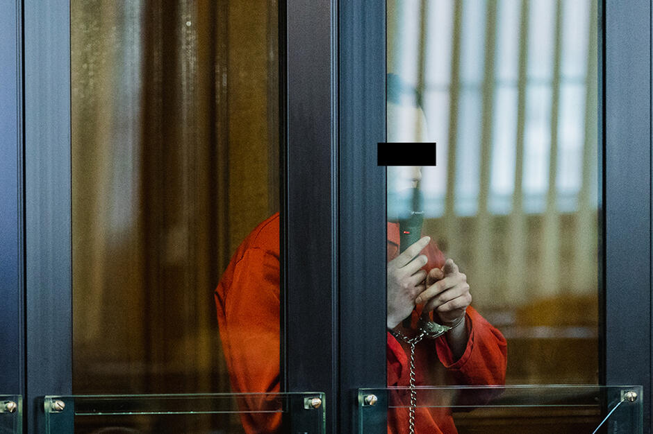 Mężczyzna w czerwonym więziennym drelichu stoi w przeszklonej klatce ochronnej dla więźniów niebezpiecznych w sali rozpraw gdańskiego sądu.
