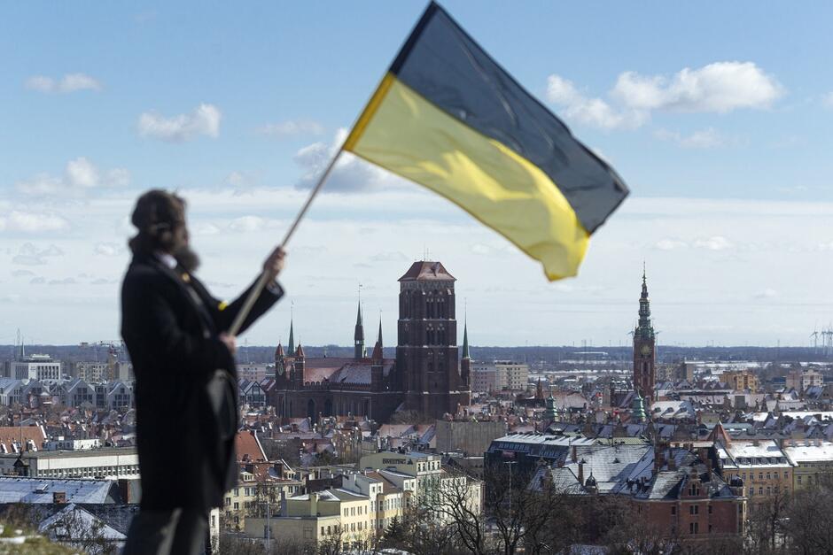 na zdjęciu mężczyzna w czarnym stroju, w tym płaszczu do kolan, trzyma w rękach czarno żółtą flagę kaszubską, którą powiewa na wzgórzu, w tle widać kamienice i zabytkowe budynki głównego miasta gdańska