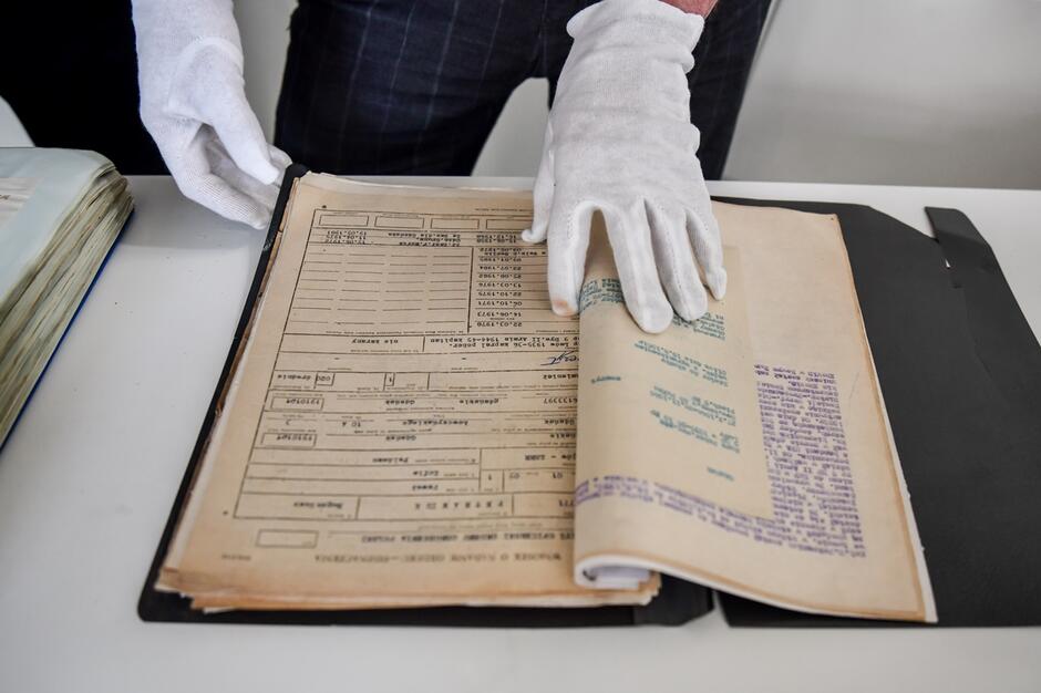 na zdjęciu stara księga, widać dwie ludzkie ręce w białych rękawiczkach, które przytrzymują księgę