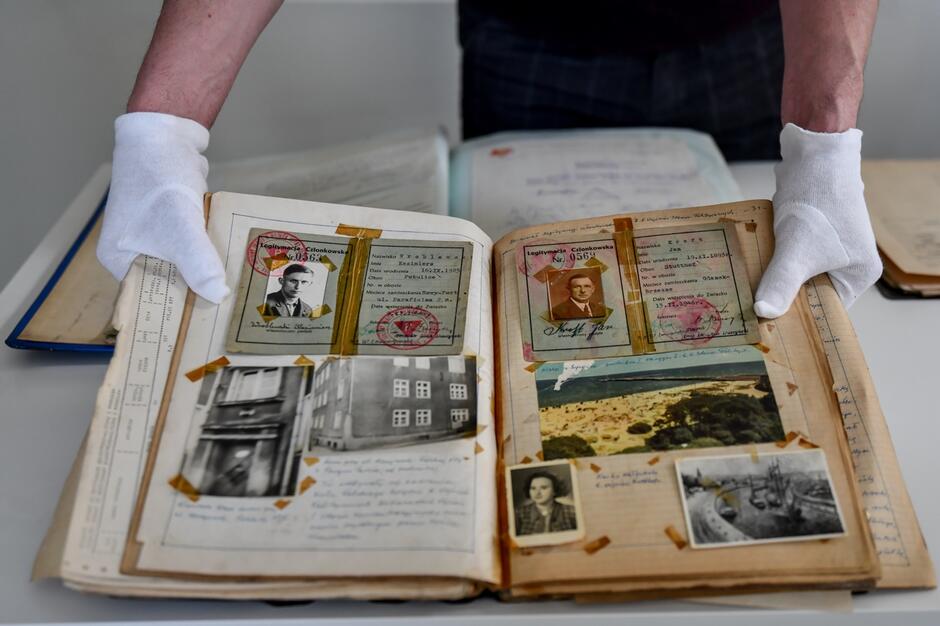 na zdjęciu stara księga z dokumentami i starymi zdjęciami, otwarta, trzymają ją dwie ludzkie ręce w białych rękawiczkach
