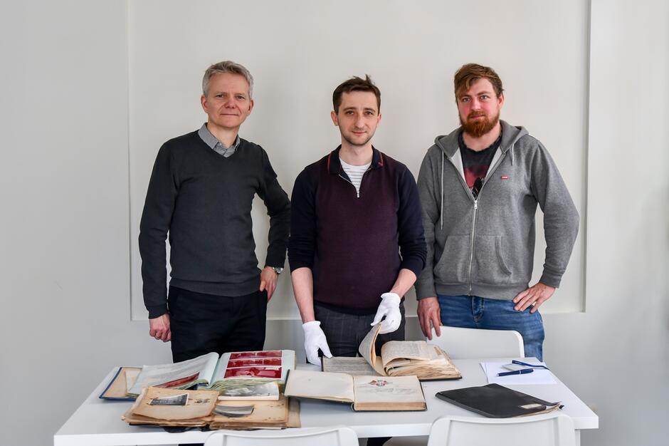 na zdjęciu trzech mężczyzn w średnim wieku, stoją przed niewielkim stolikiem na którym rozłożone są różne dokumenty