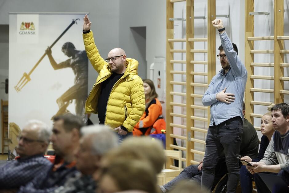 na zdjęciu dwóch mężczyzn stoi i podnosi ręce zgłaszając chęć zabrania głosu, jeden ma na sobie żółtą zimową kurtkę, drugi ma szarą koszulę, przed nimi grupa osób siedzi na krzesłach