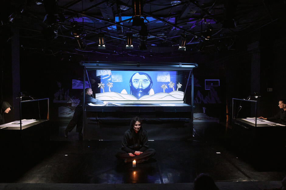 czarna sala, na podłodzie siedzi aktor, w tle niewielka zabudowana scena, widok z daleka