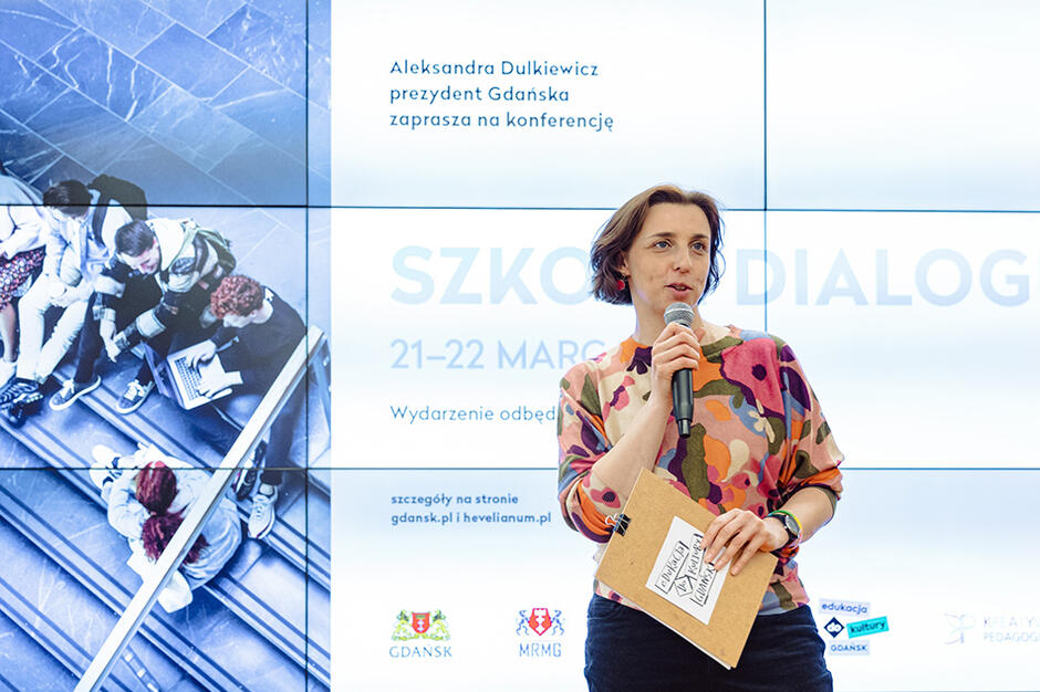 Na zdjęciu Monika Chabior, zastępczyni prezydent Gdańska ds. rozwoju społecznego i równego traktowania, mówi do mikrofonu, za nią wyświetla się wielki telebim