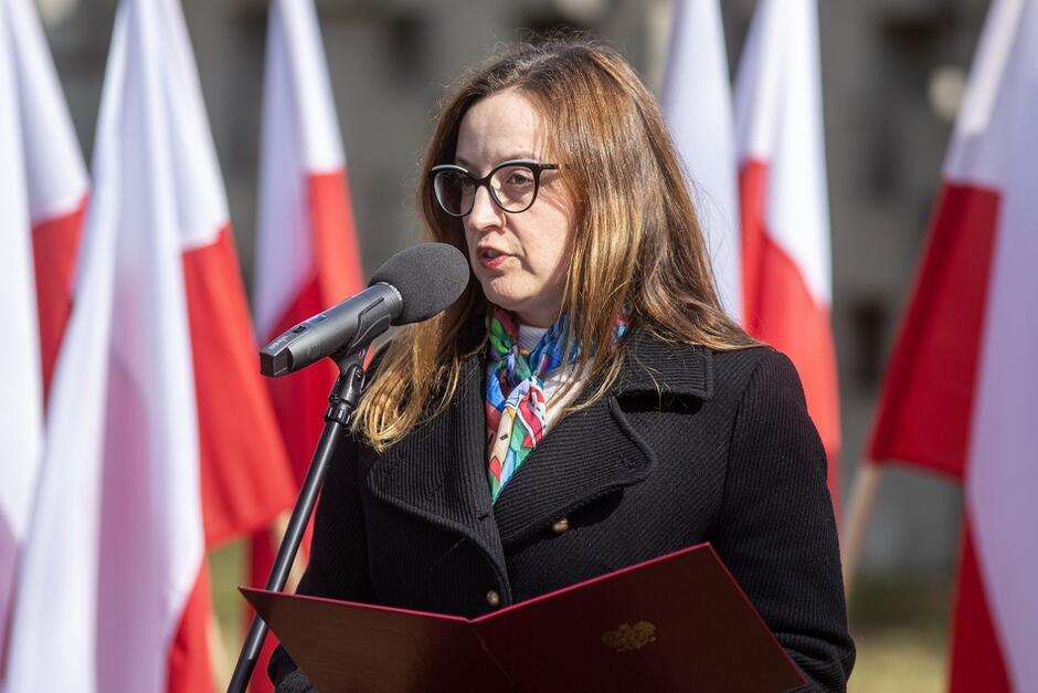 Elegancko ubrana kobieta w okularach przemawia do mikrofonu stojąc na tle biało-czerwonych flag