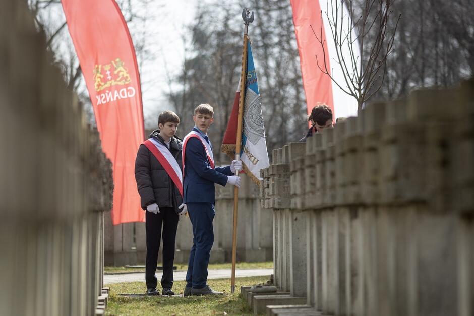 dwóch młodych chłopaków trzyma sztandar stojąc pomiędzy grobami. W tle widoczne flagi biało-czerwone oraz czerwone z herbem Gdańska.