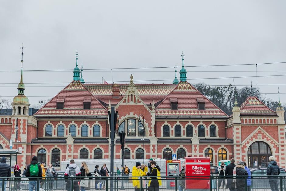 na zdjęciu jeden z budynków dworca w śródmieściu, widać jego zabytkową ceglaną elewację i dużo okien, przed budynkiem widać przystanek tramwajowy na którym stoi sporo ludzi