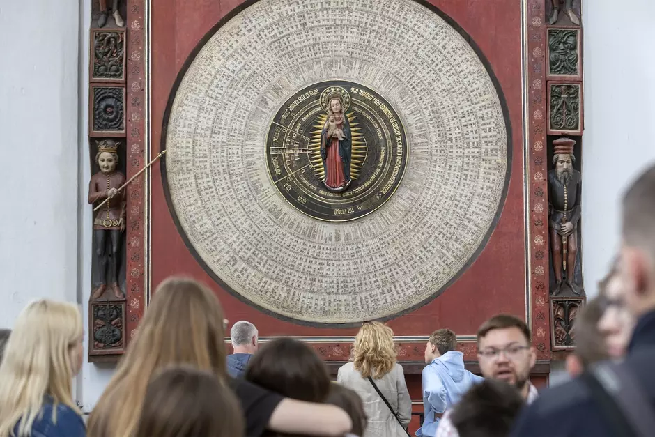 ogromna drewniana tarcza zegara na ścianie świątyni, wokół sylwetki ludzi