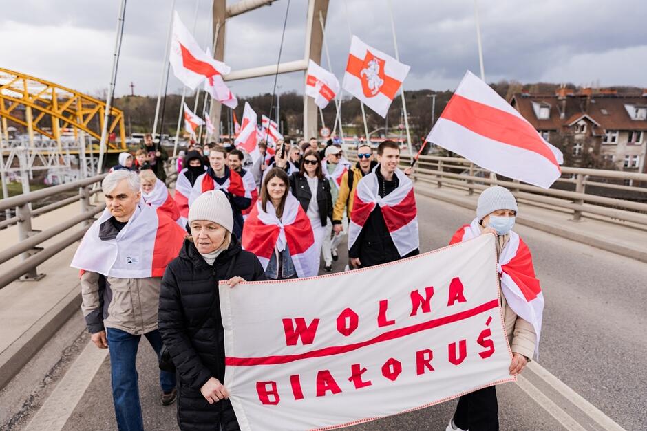 Zdjęcie przedstawia czoło pochodu. Osoby w różnym wieku, mężczyźni i kobiety, niosą biało-czerwono-białe flagi. Dwie kobiety na przedzie niosą biały prostokątny kawałek płótna z wykonanym ozdobnie na czerwono napisem Wolna Białoruś 