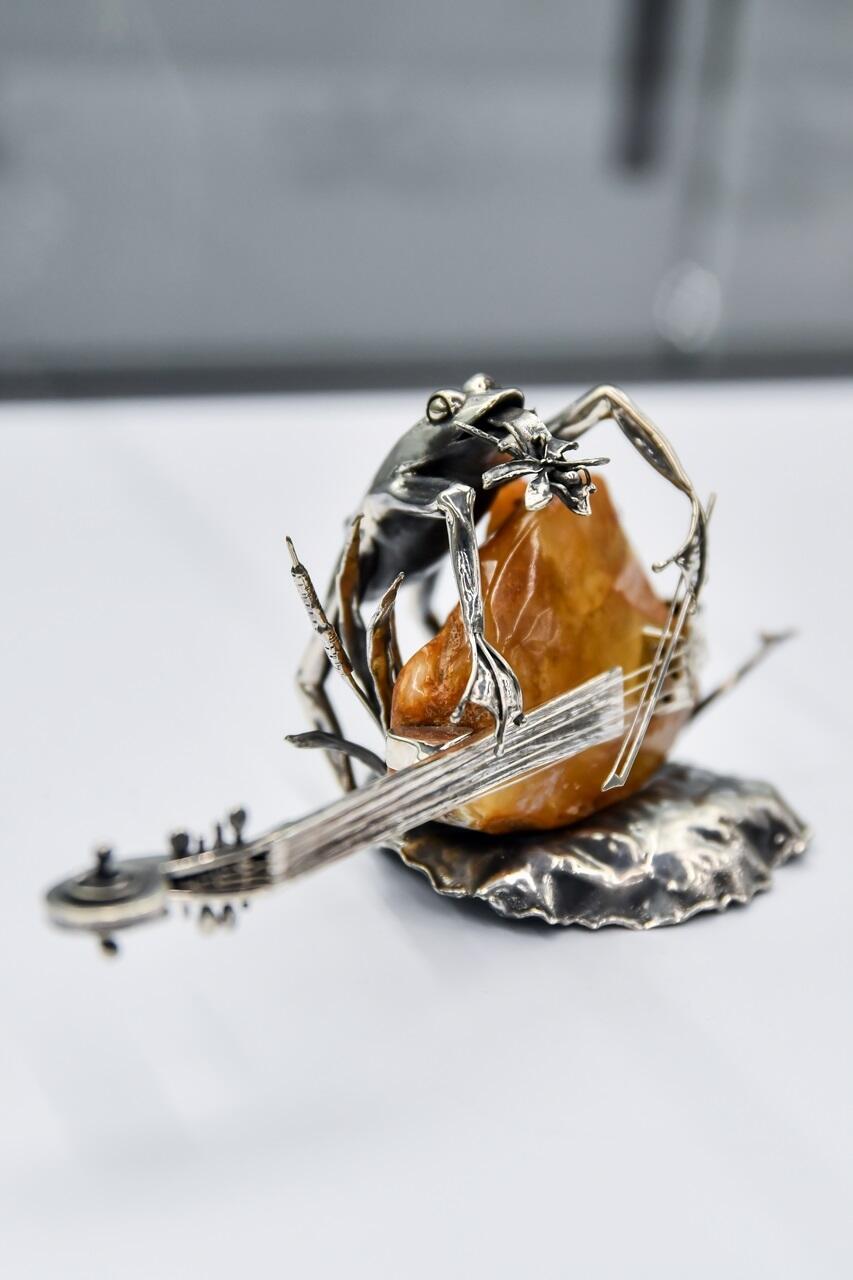 Na zdjęciu srebrna figurka żaby grającej na poziomo ułożonym kontrabasie. Korpus instrumentu wykonany jest z ciemno-miodowej bryłki bursztynu