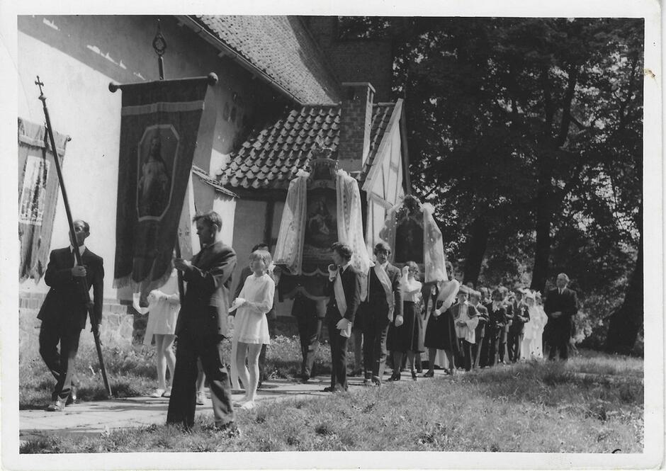 Czarno-białe zdjęcie: uczestnicy procesji idą wzdłuż białego budynku, najprawdopodobniej kościoła