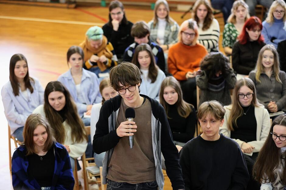 nastolatek stoi i trzyma mikrofon, wokół siedzą inni młodzi ludzie
