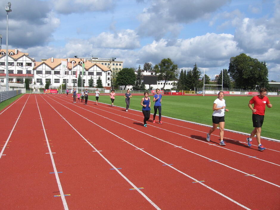 Dwie osoby biegną po pomarańczowej bieżni stadionu, bardziej po lewej (trochę z oddali) kolejne dwie osoby biegną, potem kolejne