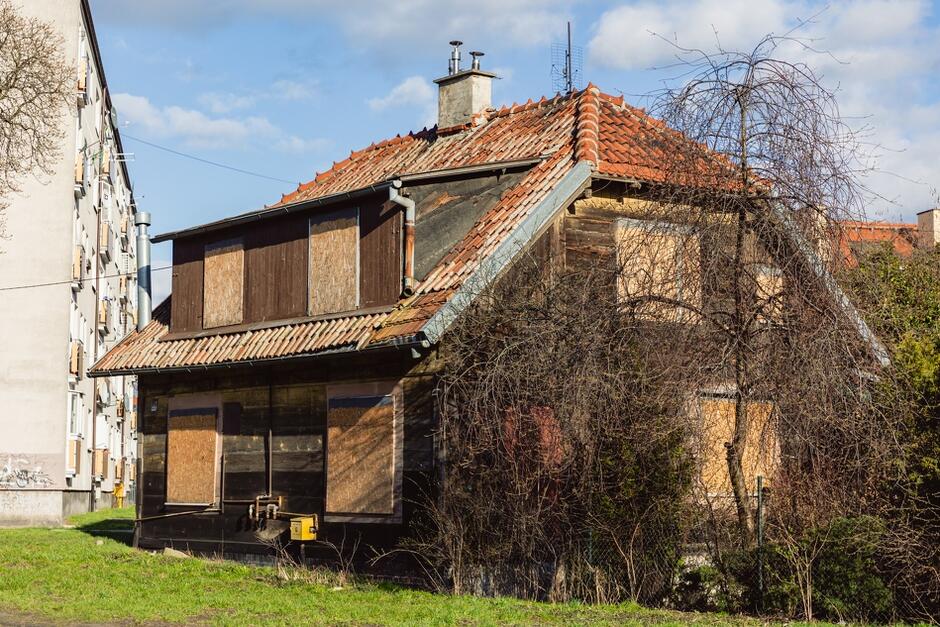 na zdjęciu stara drewniana chata dwupoziomowa, widać okna zabite deskami pilśniowymi
