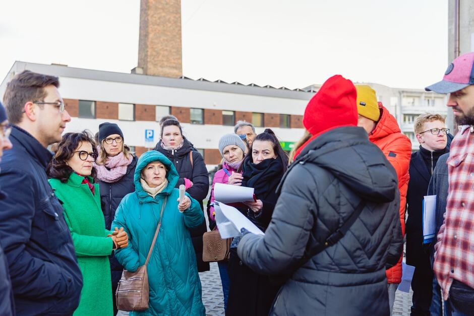 na zdjęciu grupa osób, stoją i patrzą na jedną z kobiet, która prawdopodobnie mówi, jest odwrócona plecami do fotografującego, wszyscy mają na sobie ciepłe zimowe ubrania, w różnych kolorach