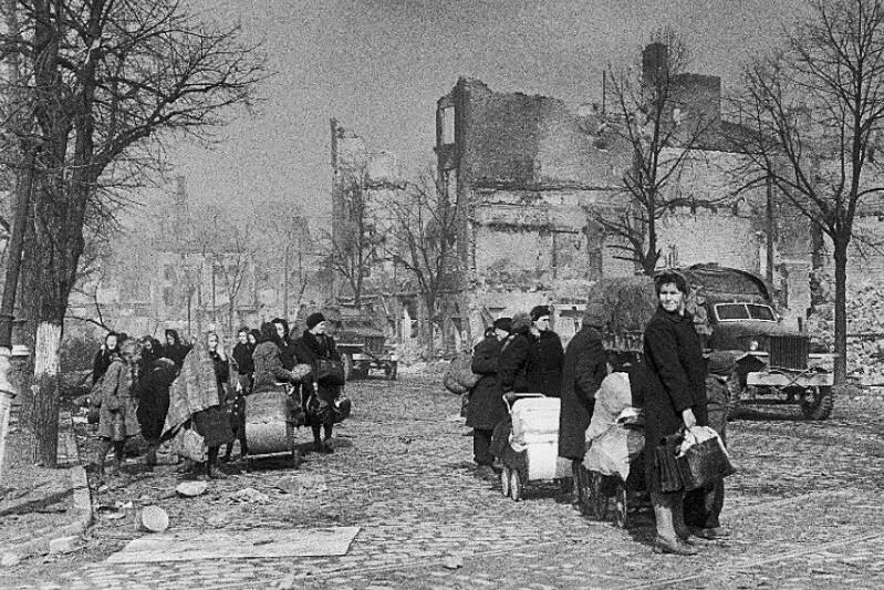Czarno-białe zdjęcie: brukowana droga, na niej ludzie z tobołkami. Po lewej drzewo, po prawej samochód, w tle budynki piętrowe
