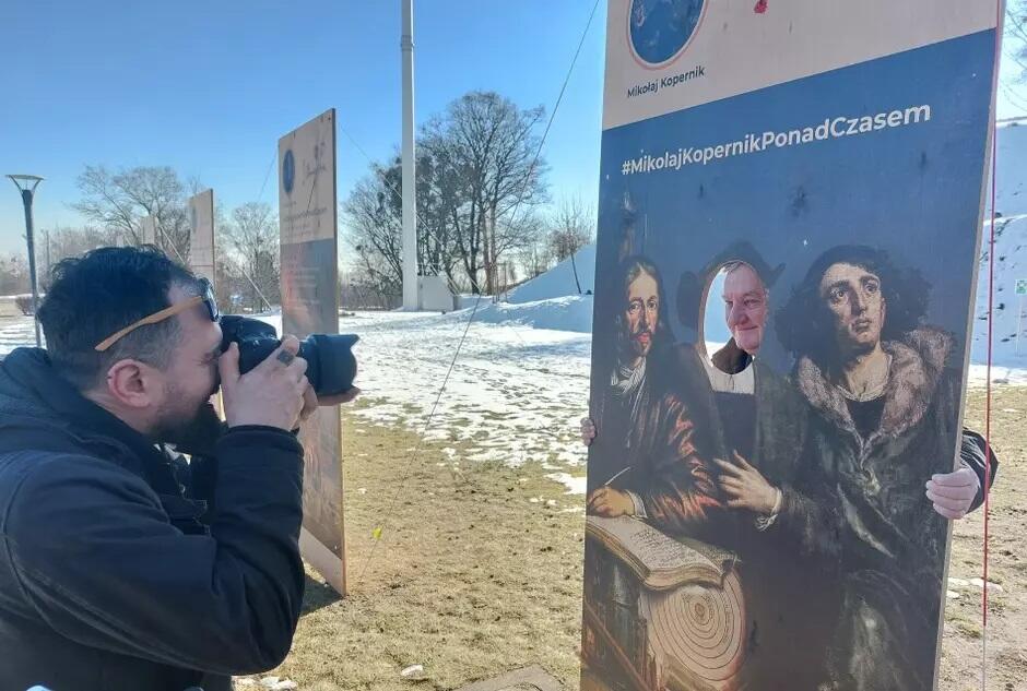 Z lewej strony młody mężczyzna z aparatem fotograficznym zbliżonym do twarzy, po prawej plansza z napisem Mikołaj Kopernik ponad czasem