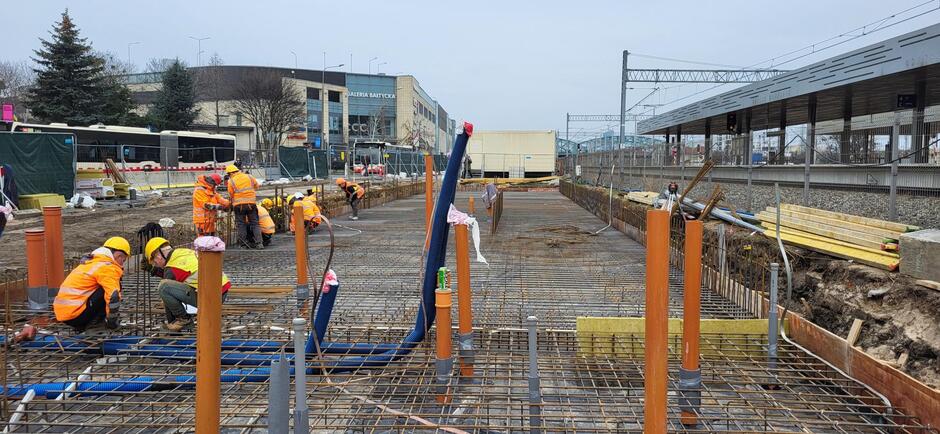 na zdjęciu plac budowy, widać kilku robotników w pomarańczowych kombinezonach którzy prowadzą prace na płycie fundamentowej