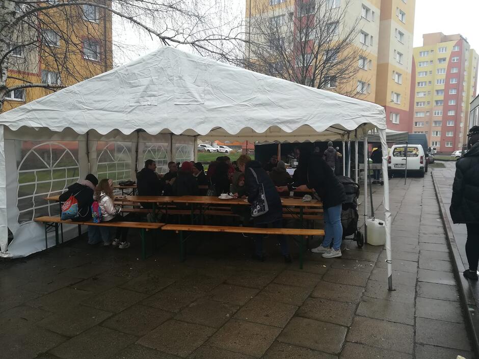 na zdjęciu duży jasny plenerowy namiot, pod nim ustawione stoły i ławy, na których siedzą ludzie
