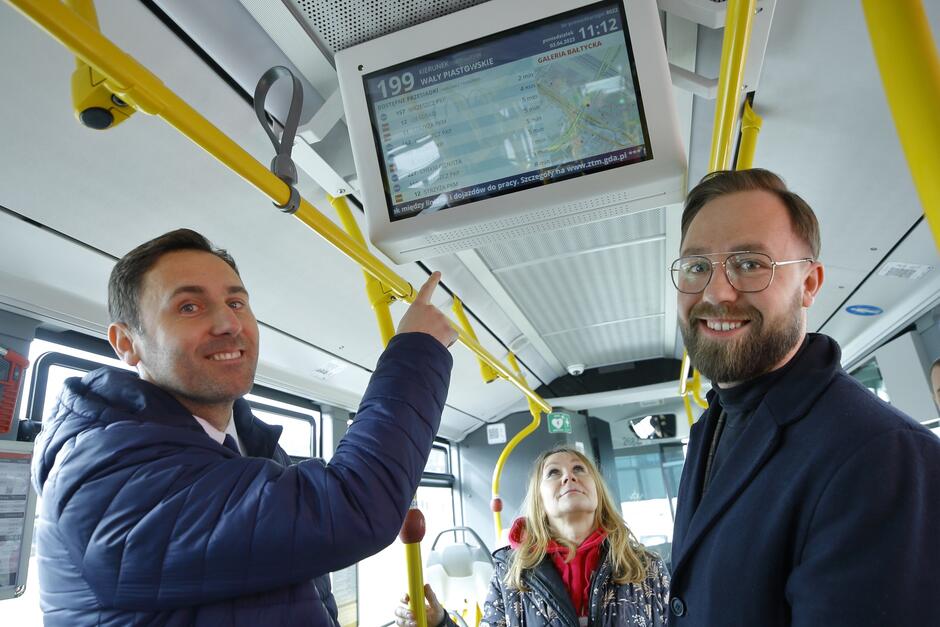 Trzy osoby w autobusie, mężczyzna po lewej wskazuje palcem na elektroniczną tablicę wiszącą wyżej 