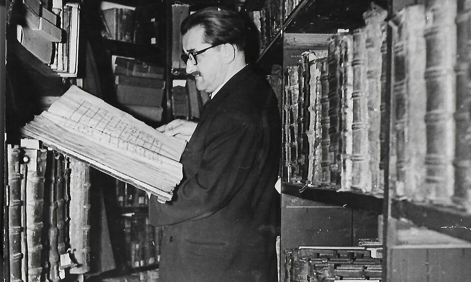 Czarno-białe zdjęcie przedstawiające mężczyznę w okularach przeglądającego książkę w bibliotece