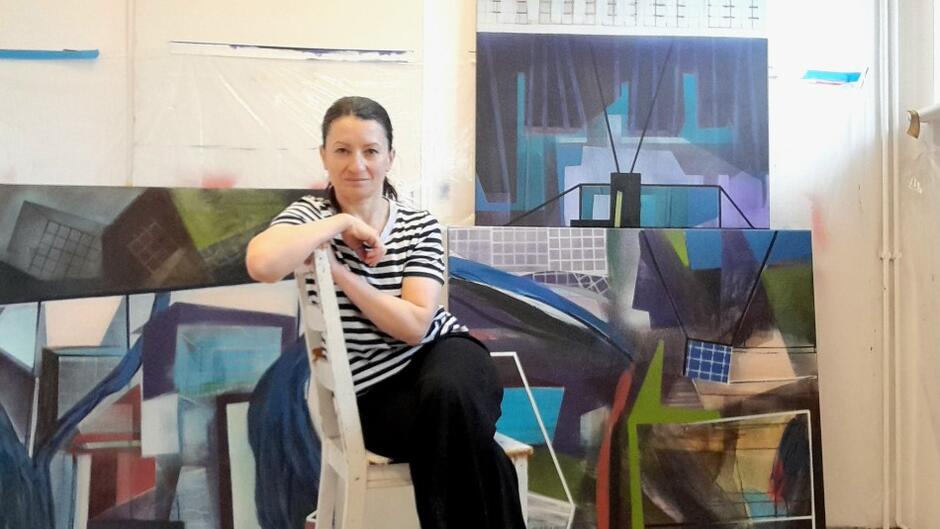 kobieta siedzi na krześle, opiera się ramionami o abstrakcyjny obraz, za nią stoją również obrazy oparte o ścianę 