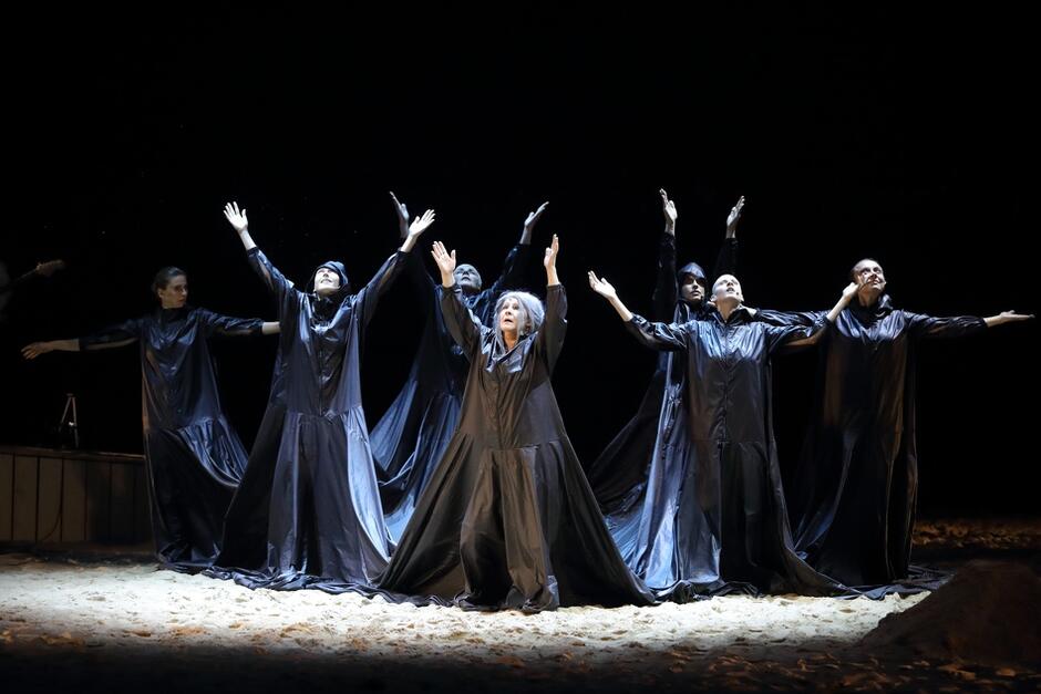 7 kobiet na scenie unosi ręce do góry, ubrane są na czarno w jedną zszytą razem szatę