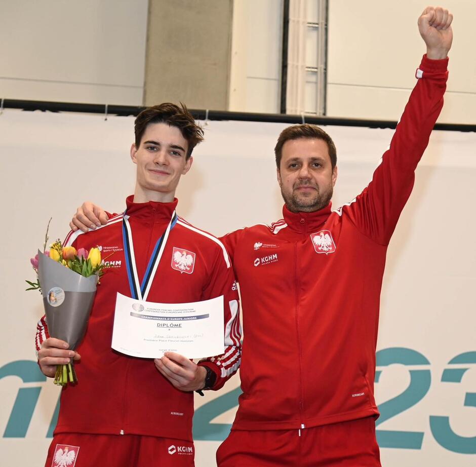 Dwóch mężczyzn w czerwonych dresach: po lewej młody z kwiatami w ręku i dyplomem, na szyi medal, po prawej starszy z wyciągniętą do góry ręką