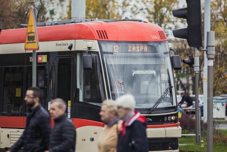 Czerwony tramwaj, na pierwszym planie pasażerowie przechodzą przez ulicę