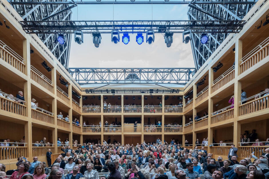 publiczność w teatrze, nad nią otwarty dach, wnętrze teatru wyłożone jasnym drewnem