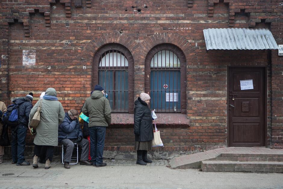 na zdjęciu kilka osób w zimowych ubraniach stoi w kolejce przed ceglanym niskim budynkiem