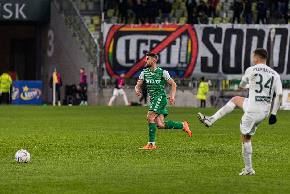 Na pierwszym planie widać piłkarzy uczestniczących w meczu. W tle jest początkowy fragment transparentu z symbolem przekreślonego telewizora z napisem LGBT na ekranie oraz pierwszymi literami napisu: SODOM