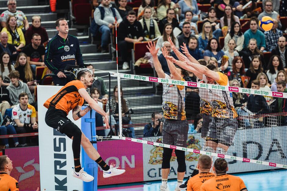 Po lewej w wyskoku atakuje wyskakuje siatkarz w pomarańczowej koszulce, po prawej za siatką dwóch zawodników w białych koszulkach z wyciągniętymi do góry rękoma. W tle trybuna z publicznością