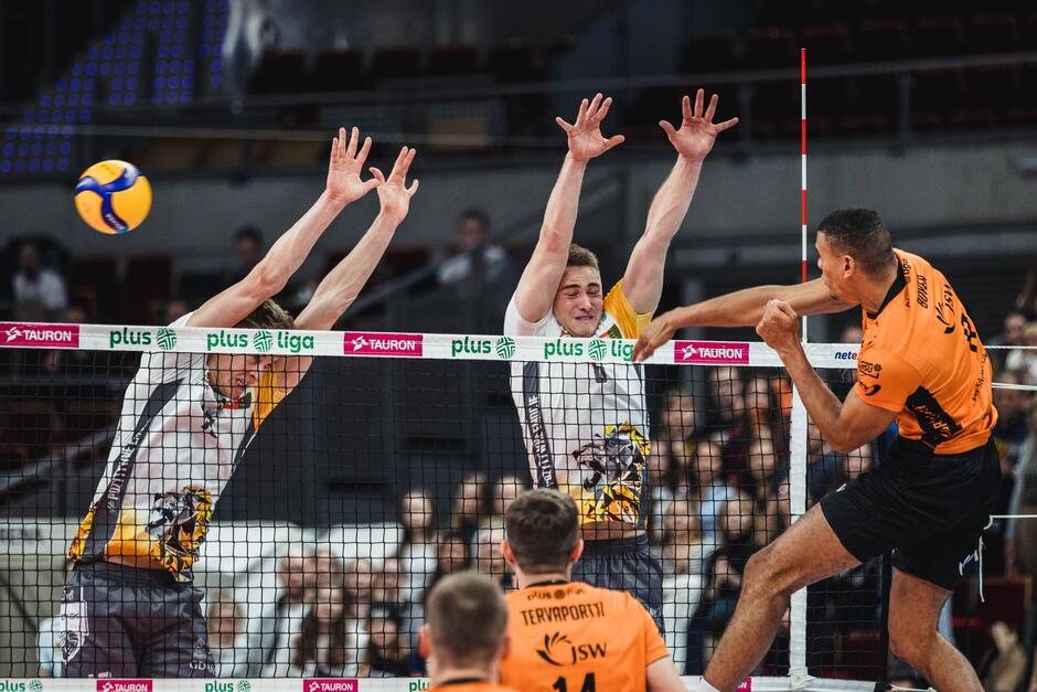Po prawej atakuje siatkarz w pomarańczowej koszulce, po lewej dwóch w wyskoku w białych koszulkach próbuje zablokować piłkę. Piłka przelatuje obok nich