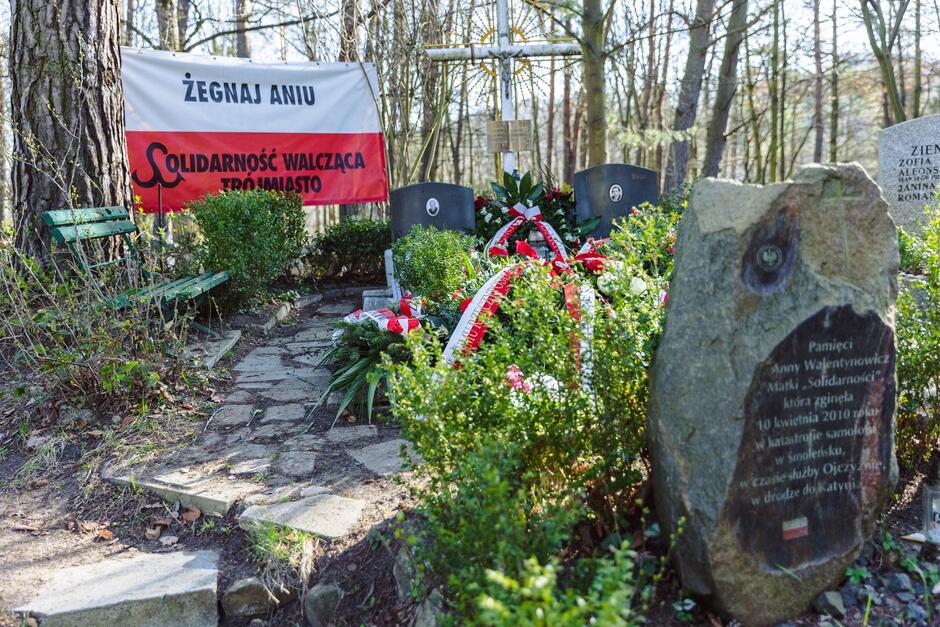 Grób wśród kwiatów i zieleni. U góry biało-czerwona flaga z napisem: ŻEGNAJ ANIU, SOLIDARNOŚĆ WALCZĄCA TRÓJMIASTO