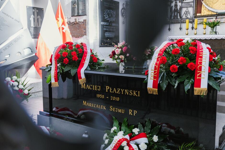 Grób w kształcie trumny w pomieszczeniu, na boku napis: Maciej Płażyński marszałek sejmu RP, po lewej flagi Polski i Gdańska. Na grobie dwa duże wieńce z biało-czerwonymi kwiatami i szarfami