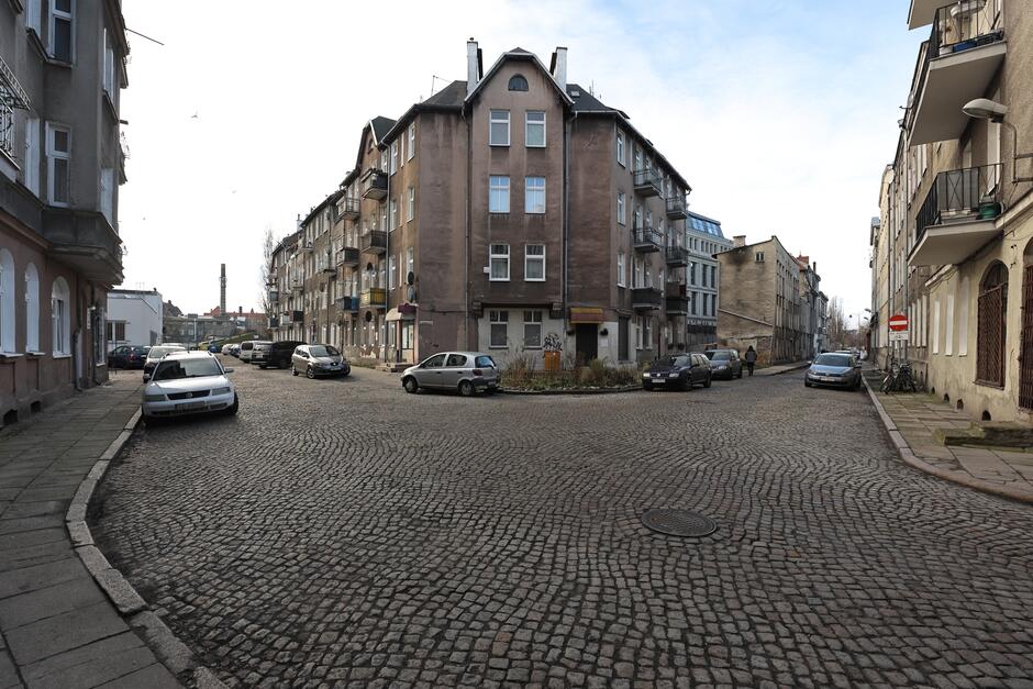 na zdjęciu fragment brukowanej ulicy, widać też szare brzydkie chodniki, w centralnym punkcie zdjęcia stoi stara kamienica o ciemnej brązowej elewacji