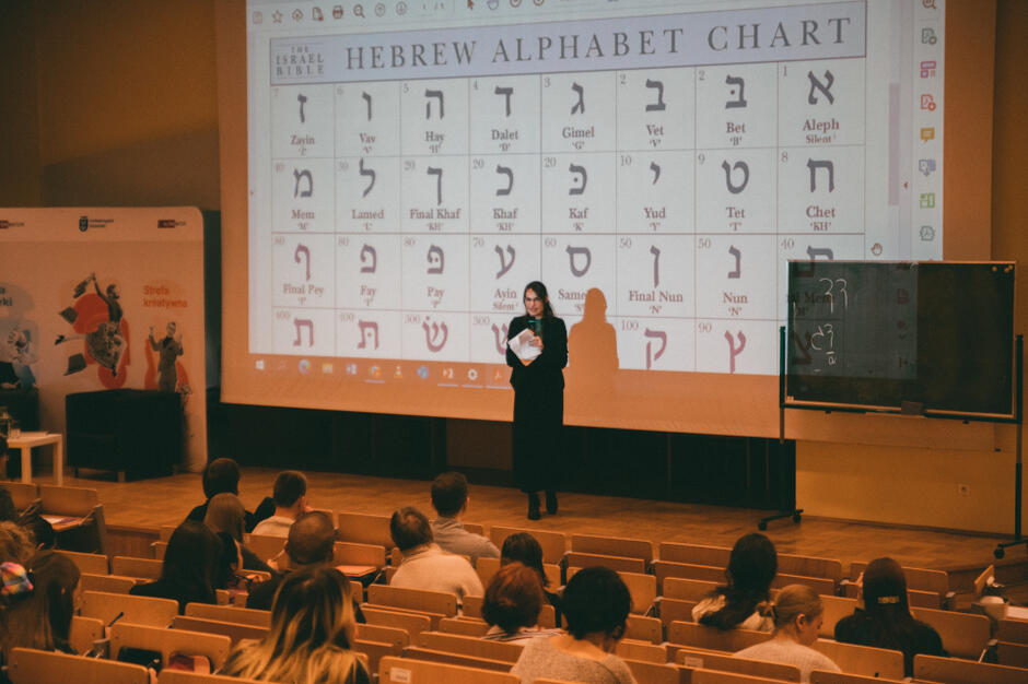 alfabet hebrajski wyświetlany na ekranie z projektora, na jego tle stoi kobieta