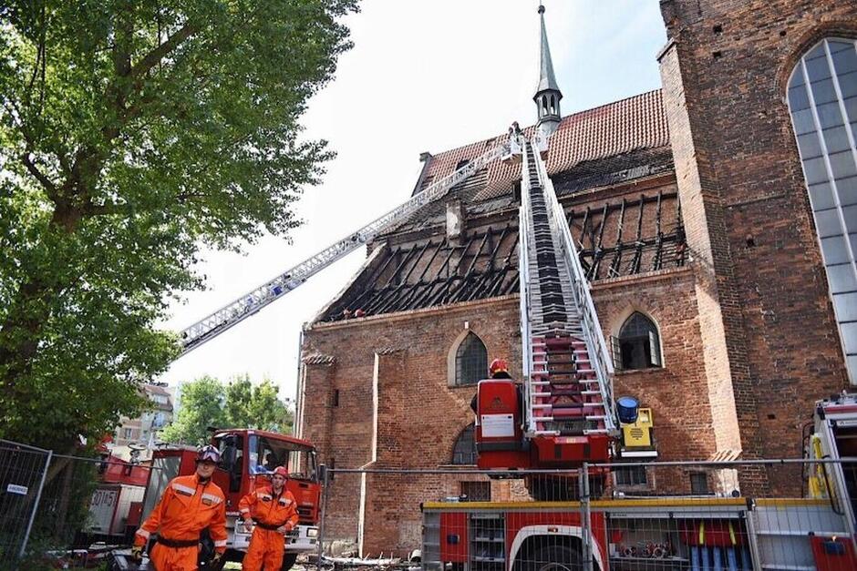 duży, ceglany kościół, na pierwszym planie strażacy i wysunięte na dach drabiny