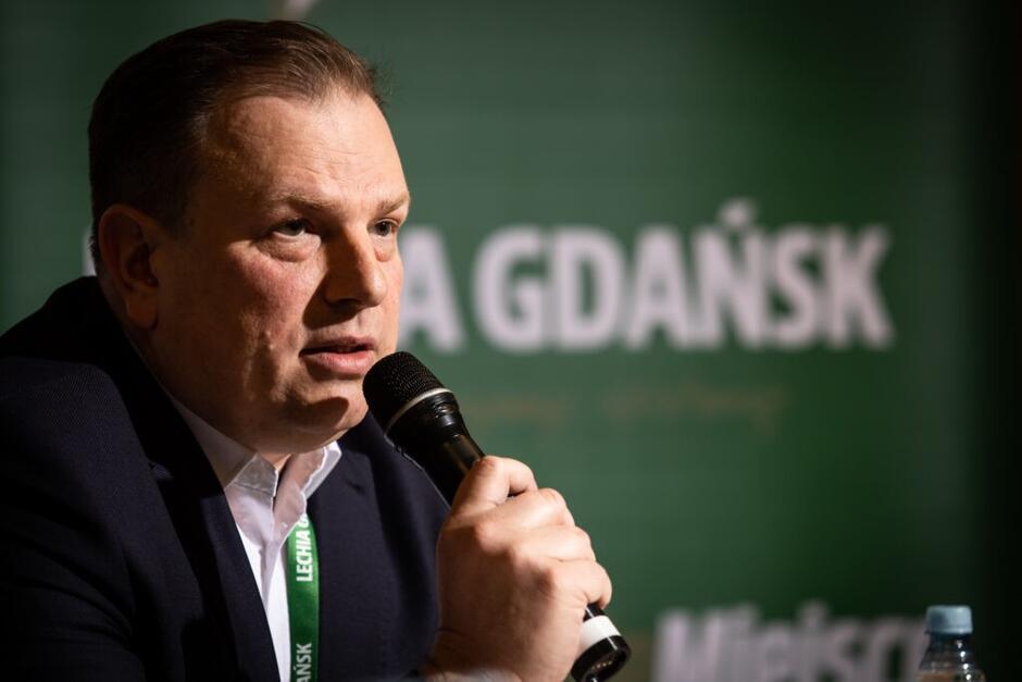 Mężczyzna w średnim wieku siedzi za stołem, mówi przez mikrofon, który trzyma w dłoni. W tle - zielona ścianka i biały napis Lechia Gdańsk