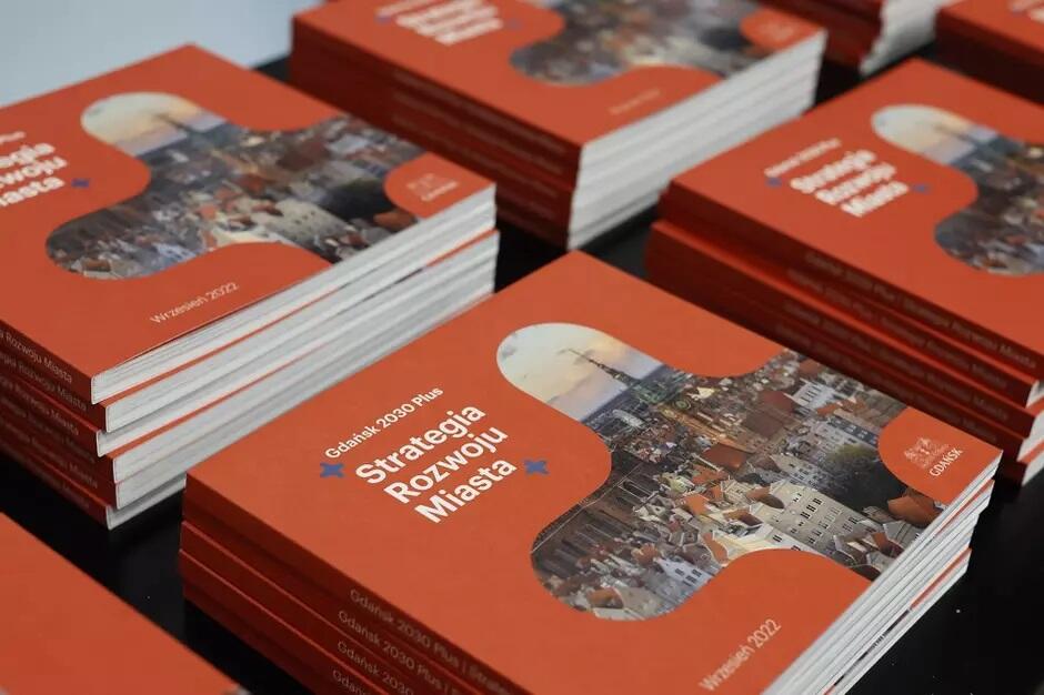 Książki o takich samych okładkach leżą obok siebie na stole. Na pomarańczowych okładkach widnieją tytuły: „Gdańsk 2030 Plus Strategia Rozwoju Miasta” 