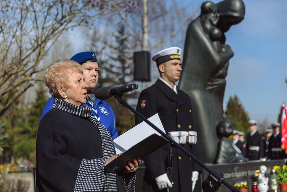 na zdjęciu starsza kobieta, mówi do mikrofonu, czytając z kartki, za nią w tle stoi młoda harcerka i żołnierz, obok stoi pomnik golgoty wschodu