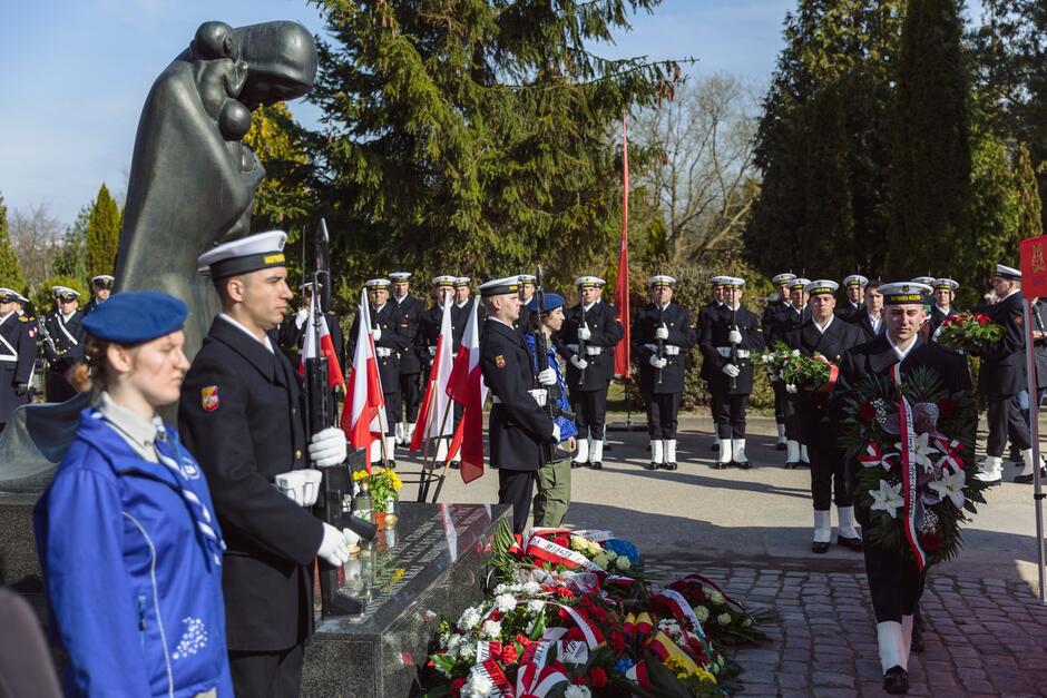 na zdjęciu żołnierz w galowym stroju idzie z wiązanką kwiatów pod pomnik, przy pomniku wartę pełni dwóch innych żołnierzy oraz harcerka, w tle widać innych żołnierzy marynarki wojennej w galowych strojach