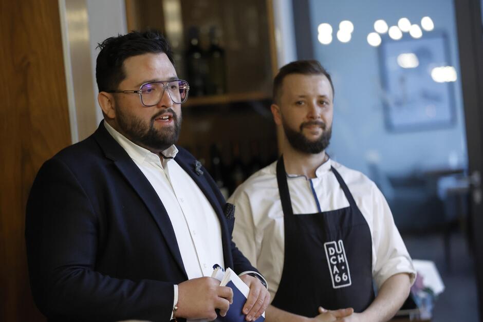 Dwaj mężczyźni stoją w restauracji. Po lewej nieco tęgi brunet w garniturze i okularach. Po prawej - szczupły brunet w fartuchu kucharskim. Obaj w wieku trzydziestu kilku lat 