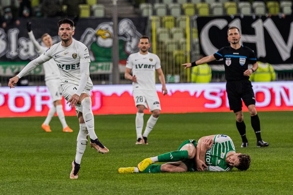 Piłkarz w biało-zielonym stroju leży na murawie, trzyma się za rękę. Za nim mężczyzna w czarnym stroju, obok po lewej trzech piłkarzy w białych strojach