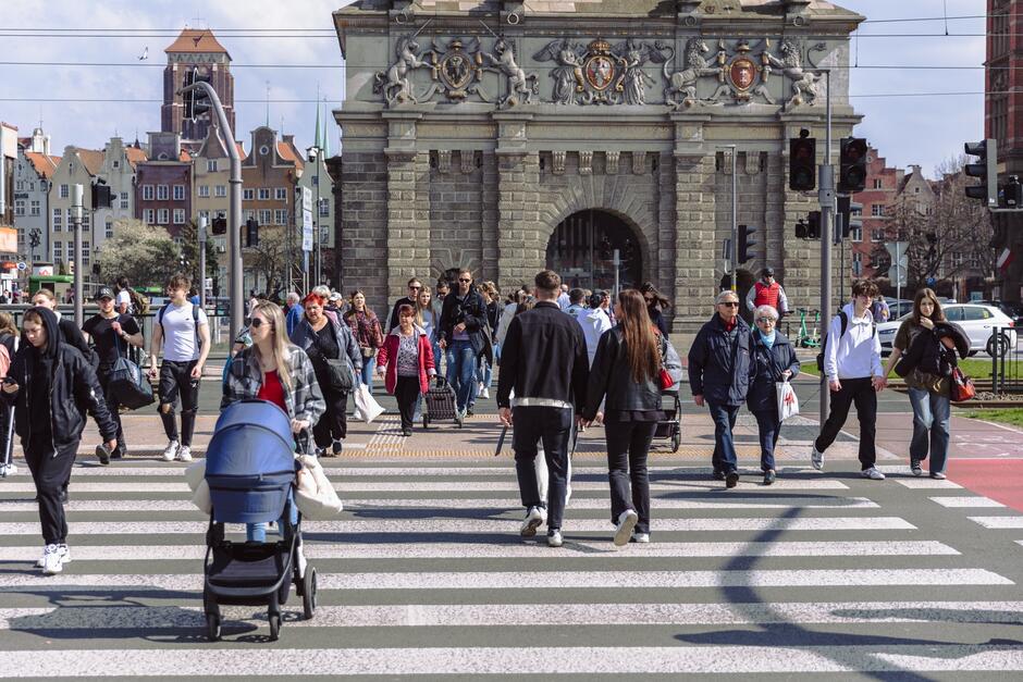 na zdjęciu przejście naziemne dla pieszych, po którym przechodzi duża grupa osób, w tle widać kilka zabytkowych budynków w centrum gdańska, w tym zabytkową bramę wyżynną