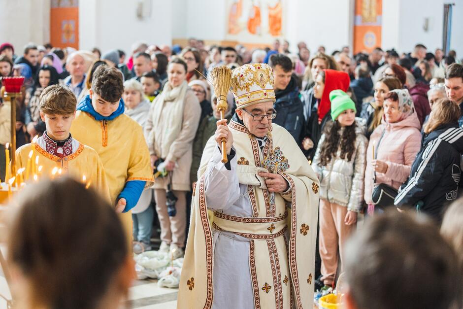 Wnętrze świątyni. Kapłan w stroju prawosławnym macha kropidłem na stojące wokół niego osoby z koszyczkami, w których są pokarmy do święcenia. Osób wokół jest dużo, kilkadziesiąt 