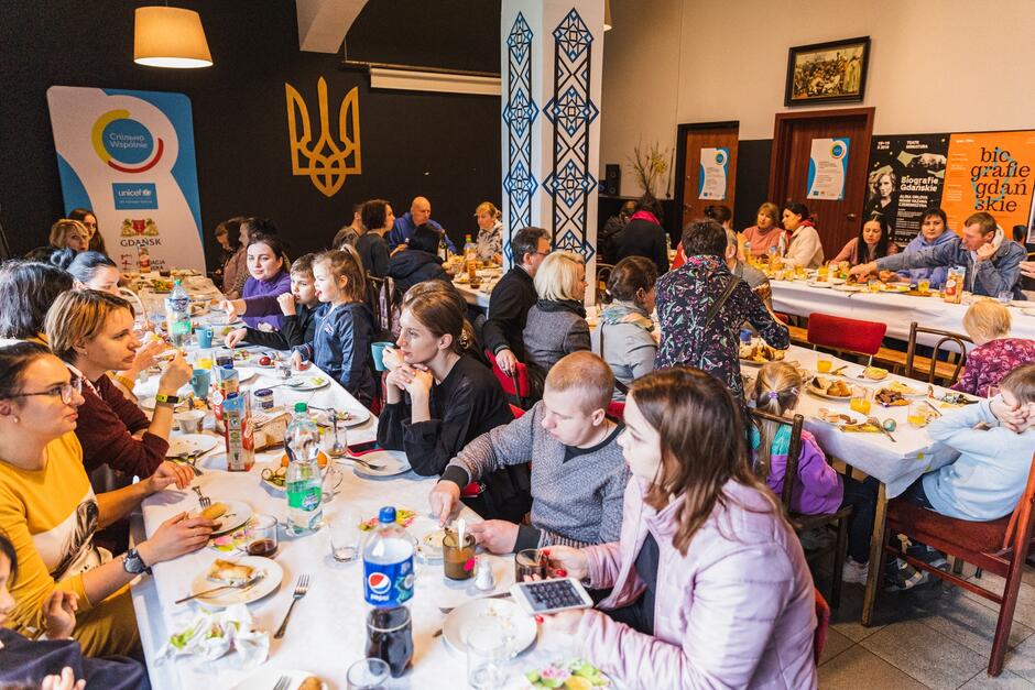 Kilkadziesiąt osób siedzi przy długich stołach zastawionych pokarmami