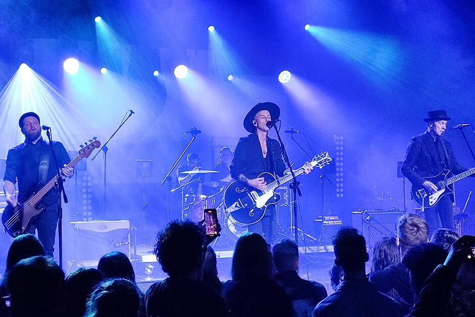Zdjęcie zespołu występującego na scenie, która jest oświetlona na niebiesko. Na pierwszym planie, w równych odstępach stoi trzech mężczyzn z gitarami, przed nimi mikrofony, do których śpiewają. Po lewej - basista z brodą, pośrodku - muzyk w kapeluszu, z gitarą, po prawej - muzyk w kapelusiku, z gitarą 